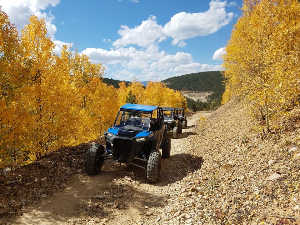 A blue Polaris RZR XP leading two more UTVs down a rocky mining road between gold aspens