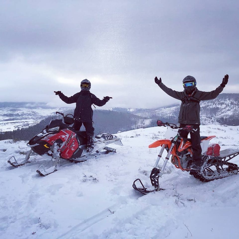 A snowmobiler and a snowbike rider standing arms-out on a snowy ridge with valleys and clouds behind