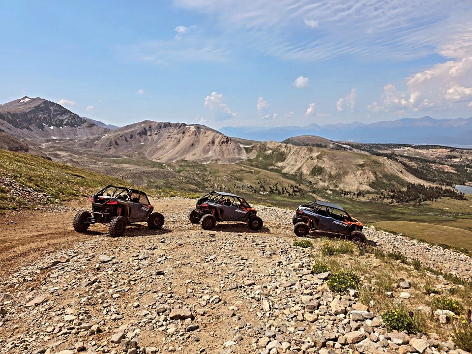 Three Polaris RZR side-by-sides parked in a row on a rocky high plateau, bare peaks and a long mountain range behind
