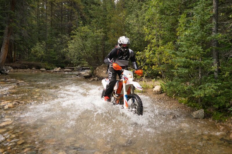 A KTM dirt bike rider splashing through a shallow creek in the forest